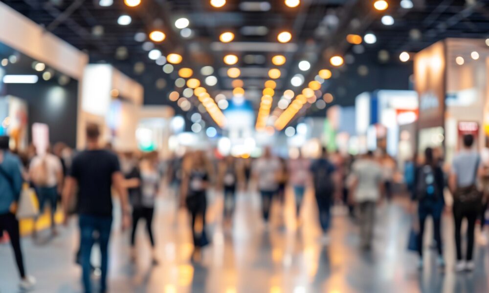 Blurry image capturing the bustling atmosphere of a trade show, with many attendees moving between booths and exhibits.