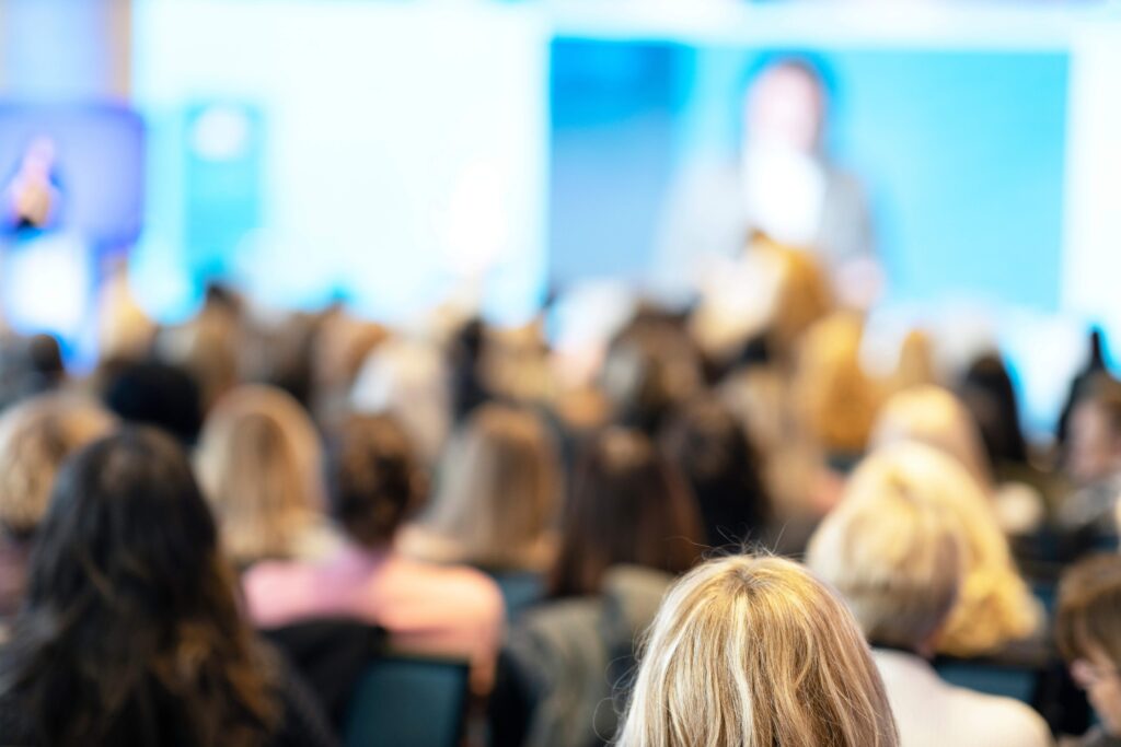Audience attending a hybrid event, with both in-person participants seated in the venue and virtual attendees shown on large screens.