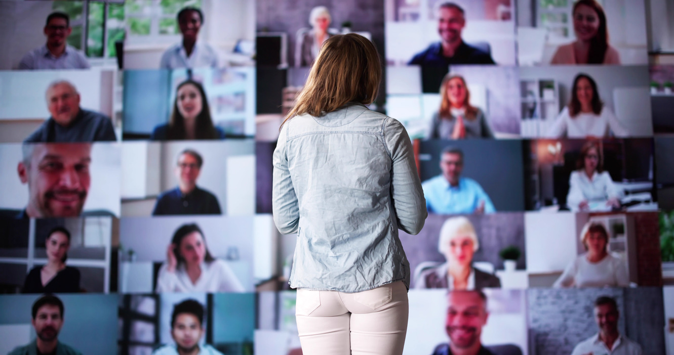 Speaker presenting to a virtual audience with multiple displays showing participants during a hybrid event.