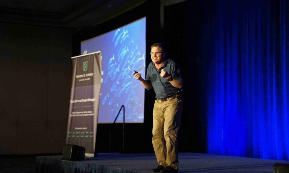 Confident business speaker standing centre stage at a leadership event, addressing an audience with strong posture and engaged eye contactConfident business speaker standing centre stage at a leadership event, addressing an audience with strong posture and engaged eye contact