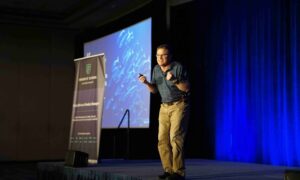 Confident business speaker standing centre stage at a leadership event, addressing an audience with strong posture and engaged eye contactConfident business speaker standing centre stage at a leadership event, addressing an audience with strong posture and engaged eye contact