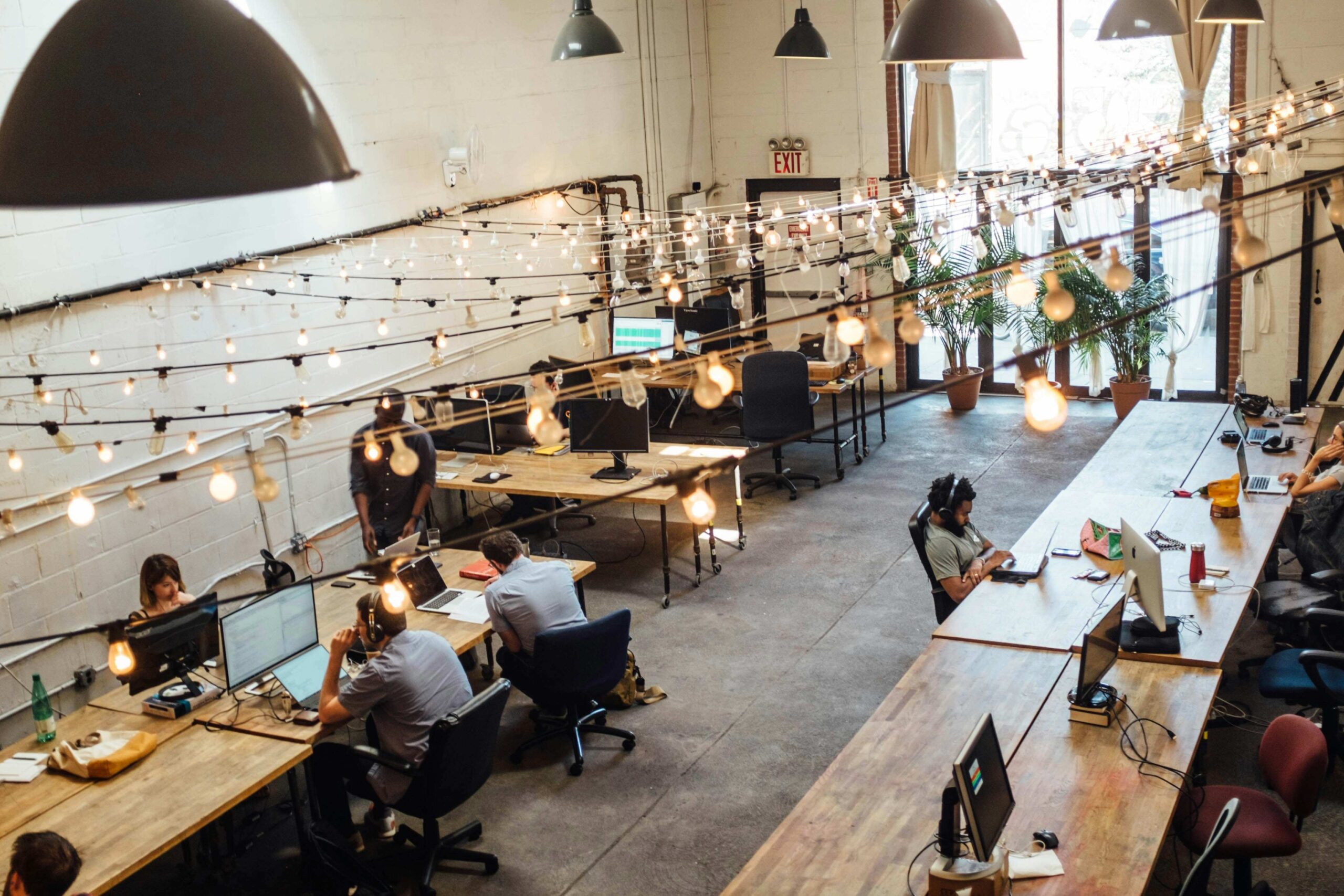Open-plan office with employees at desks attending virtual meetings on laptops and monitors, illustrating hybrid working, digital collaboration, and internal communication in a modern workplace.